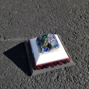 May include: A small, white pyramid with a burgundy base. The top of the pyramid is made of clear resin and contains a variety of colorful gemstones, including green, blue, and purple.