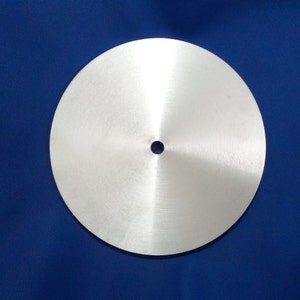 Puede incluir: Un disco circular de metal cepillado con un pequeño agujero en el centro. El disco es plateado y refleja la luz, creando un brillo sutil. El fondo es de un azul oscuro sólido.