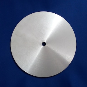 Puede incluir: Un disco redondo de metal de color plateado con un pequeño agujero en el centro. El disco tiene un acabado cepillado y es ligeramente cóncavo.