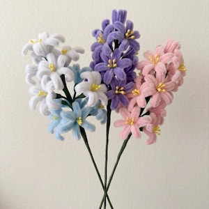 May include: A bouquet of handmade flowers in shades of white, light blue, purple, and pink. Each flower has yellow centers and is attached to a green stem. The flowers are made of a soft, fuzzy material.