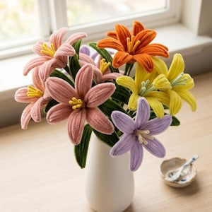 May include: A bouquet of colorful, handmade flowers in a white vase. The flowers are in shades of pink, orange, yellow, and purple, with green stems and leaves. The vase sits on a light-colored wooden surface near a window.
