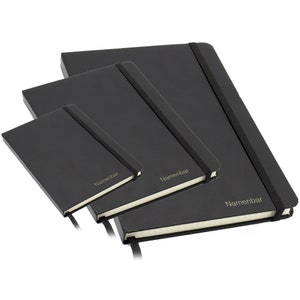 Könnte beinhalten: Drei schwarze, ledergebundene Notizbücher in verschiedenen Größen, jedes mit einem goldenen Gummibandverschluss und dem Wort "Namenbar" in Gold auf dem Einband geprägt.