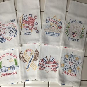 May include: Eight white tea towels with red, white, and blue embroidered patriotic designs. The designs include the words "God Bless the USA", "True to the Red, White & Blue", "Liberty", "We the People", "Proud to be an American", "Support Our Troops", "Home of the Brave", and "America".