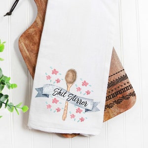 May include: White kitchen towel with a watercolor illustration of a wooden spoon, pink flowers, and a blue banner with the text "Shit Stirrer".