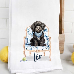 May include: A white kitchen towel featuring a black and white illustration of a dog sitting in a blue and gold chair. The dog is wearing a blue bandana and the name "Leo" is written below the chair.