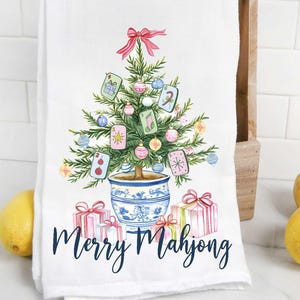 May include: White kitchen towel with a watercolor illustration of a Christmas tree decorated with Mahjong tiles, ornaments, and a red bow. The towel has the words "Merry Mahjong" in navy blue script, with wrapped gifts at the base.