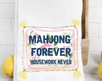 May include: White kitchen towel with the text "MAHJONG FOREVER HOUSEWORK NEVER" in navy blue, surrounded by a floral design and a gold rope border. Yellow tassels adorn the corners. Lemons are in the background.