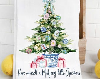 May include: White kitchen towel featuring a Christmas tree illustration adorned with Mahjong tiles and ornaments. The tree is in a blue and white pot, with wrapped gifts below. The text reads "Have yourself a Mahjong little Christmas."