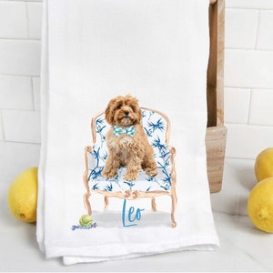 Puede incluir: Una toalla de cocina blanca con una ilustración de acuarela de un perro marrón con una pajarita azul sentado en una silla con estampado azul y blanco. El nombre del perro, "Leo", está escrito en cursiva azul debajo de la silla.
