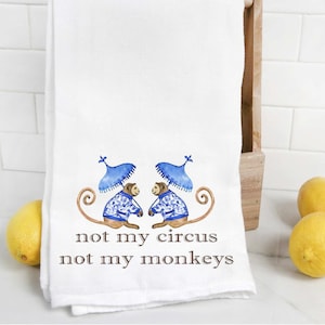 May include: White kitchen towel with two monkeys wearing blue hats and blue and white patterned clothing. The text "not my circus not my monkeys" is printed on the towel.