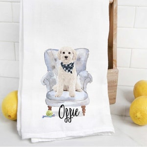 May include: White kitchen towel with a watercolor illustration of a dog sitting in a blue armchair. The dog is wearing a black and white checkered bandana. The dog's name, "Orrie", is written in black cursive below the chair.