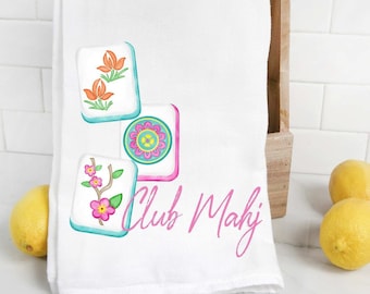 May include: White kitchen towel with a pink, blue and green design of mahjong tiles. The tiles feature flowers and a circle pattern. The text "Club Mahj" is written in pink cursive.