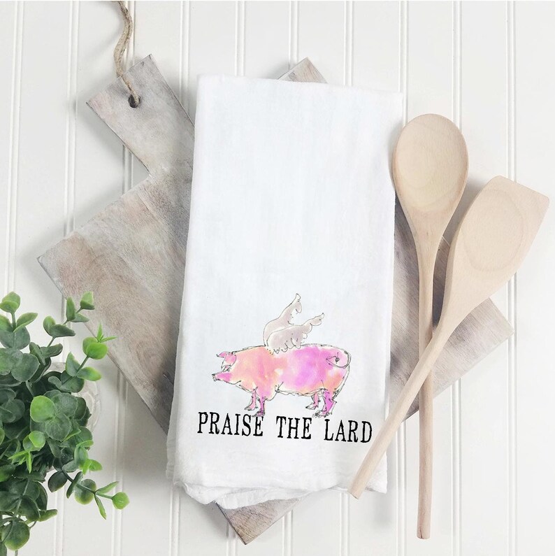 May include: White kitchen towel with a pink pig with wings and the text "PRAISE THE LARD" printed on it.