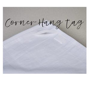 May include: White corner hang tag with the text "Corner Hang tag" in a script font. The tag is made of a textured fabric and has a small rectangular label attached.