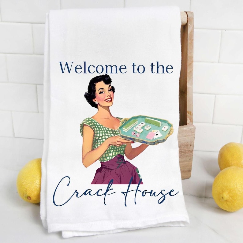 Peut inclure: Serviette de cuisine blanche avec une illustration r&eacute;tro d'une femme tenant un plateau avec des pi&egrave;ces de jeu. Le texte "Welcome to the Crack House" est &eacute;crit en bleu marine.