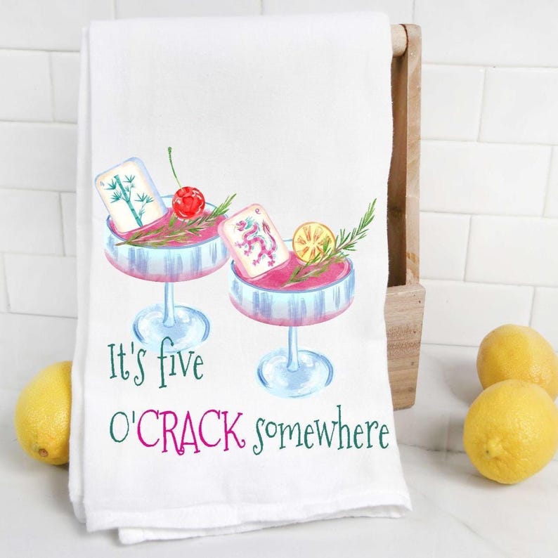 May include: White kitchen towel with a watercolor illustration of two cocktail glasses filled with pink liquid, garnishes, and playing cards. The text "It's five O'CRACK somewhere" is printed in green and pink.
