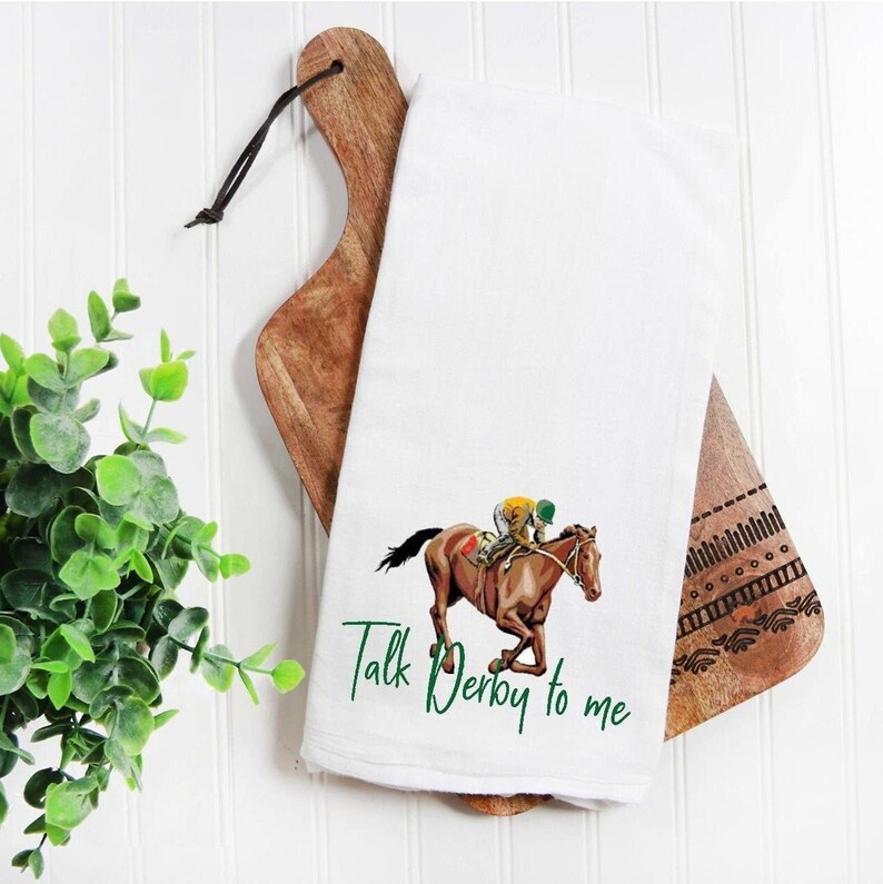 May include: White kitchen towel with a green and brown illustration of a horse and jockey. The text "Talk Derby to me" is written in green below the image.