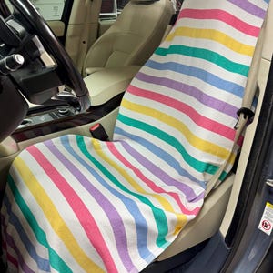 May include: A colorful striped car seat cover in a vehicle. The cover features horizontal stripes in various colors, including pink, yellow, green, purple, and blue. The cover is draped over the seat and extends down.
