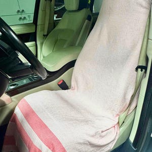 Könnte beinhalten: Ein hellrosa und weiß gestreiftes Baumwollhandtuch, das über einen Autositz drapiert ist. Das Handtuch hat ein strukturiertes Gewebe mit horizontalen rot-rosa Streifen an den oberen und unteren Kanten sowie einen gefransten Saum an der Unterseite.