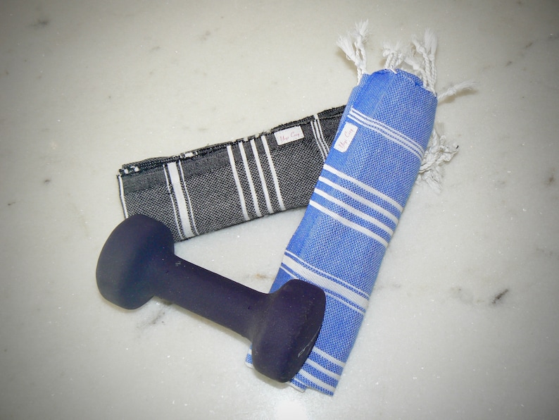 K&ouml;nnte beinhalten: Zwei t&uuml;rkische Handt&uuml;cher, eines schwarz-wei&szlig; gestreift und eines blau mit wei&szlig;en Streifen, liegen gefaltet auf einer wei&szlig;en Marmorfl&auml;che. Eine schwarze Hantel liegt auf den Handt&uuml;chern.