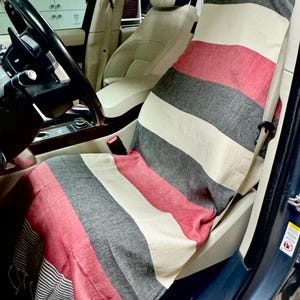 Könnte beinhalten: Ein rechteckiger Autositzbezug mit horizontalen Streifen in Rot, Anthrazit und Creme. Der Stoff hat eine gewebte Textur und eine Fransenkante am unteren Ende, drapiert über dem Beifahrersitz eines Fahrzeugs.