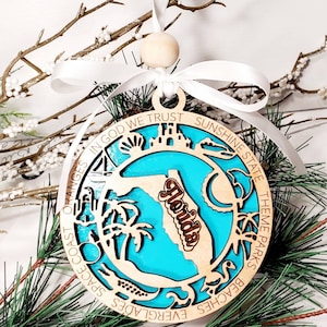 Op de afbeelding: Houten Florida staat ornament met een turquoise achtergrond en een licht houten frame. Het ornament heeft het woord "Florida" in donkerder hout en de tekst "IN GOD WE TRUST SUNSHINE STATE THEME PARKS BEACHES EVERGLADES SPACE COAST ORANGES". Het hangt aan een wit lint.