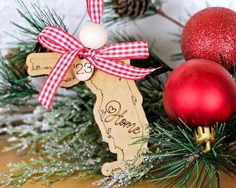 May include: A wooden Christmas ornament shaped like the state of Florida with the words "Home" and a heart inside the state outline. The ornament has a red and white checkered ribbon and a wooden bead. The ornament is surrounded by pine branches and red Christmas ornaments.