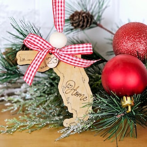 Könnte beinhalten: Ein hölzerner Weihnachtsbaumschmuck in Form des Bundesstaates Florida mit den Worten "Home" und einem Herzen innerhalb der Staatsgrenze. Der Schmuck ist mit einem rot-weiß karierten Band und einer Holzperle verziert. Der Schmuck ist von Kiefernästen und roten Weihnachtskugeln umgeben.