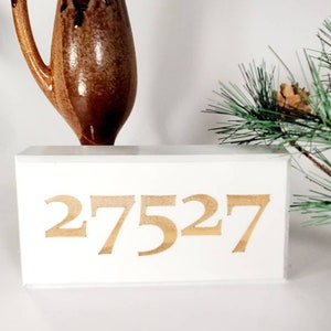 May include: White wooden block with the number 27527 printed in brown. The block is sitting on a white surface next to a brown vase and a green pine branch with brown pine cones.