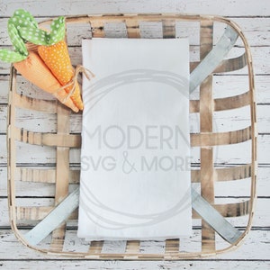 May include: A white folded towel rests in a rustic, square woven basket. Two orange fabric carrots with green polka dot tops are tied together with twine. The basket is set against a white wooden background.