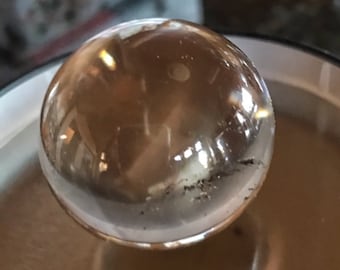 May include: A clear glass sphere with a slightly cloudy appearance. The sphere is reflecting light and has a few small dark spots on the surface.