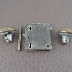May include: An antique door lock set, comprising a rectangular lock body, two rounded knobs, and a threaded rod. The metal components display signs of age and wear, with silver and brown tones.