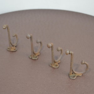 May include: Four antique metal hooks with a curved design, arranged on a brown surface. The hooks have a weathered, aged appearance, suggesting they are vintage or have been exposed to the elements. They are likely used for hanging items.