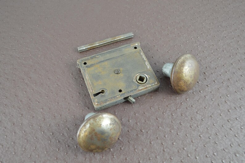 May include: A vintage door lock set featuring two oval brass doorknobs, a square lock plate, and a threaded rod. The metal components have a tarnished, aged appearance, suggesting vintage hardware. The set is displayed on a textured brown surface.