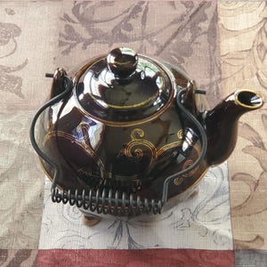 May include: A brown ceramic teapot with a decorative gold swirl pattern. The teapot has a black metal handle and a spout with a gold rim. The lid has a small, round knob. The teapot is sitting on a patterned surface.