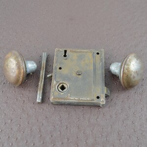 May include: An antique door lock set featuring two round doorknobs and a rectangular lock mechanism. The metal components have a tarnished, aged appearance, suggesting vintage hardware. The set includes a spindle and is likely for a door.