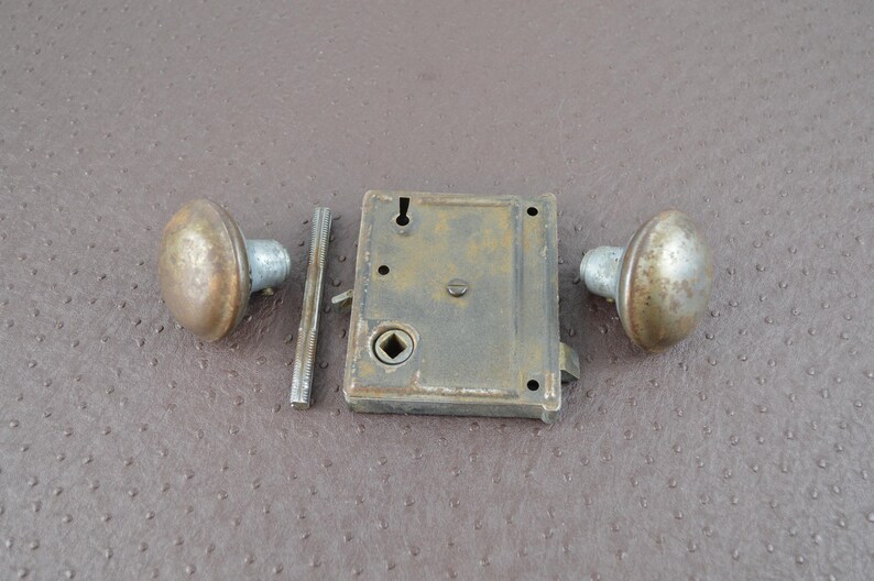 May include: An antique door lock set featuring two round doorknobs and a rectangular lock mechanism. The metal components have a tarnished, aged appearance, suggesting vintage hardware. The set includes a spindle and is likely for a door.
