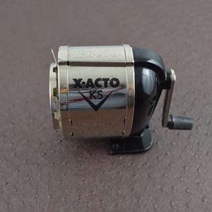 May include: A silver and black X-ACTO KS pencil sharpener. The sharpener has a crank handle and a silver blade guard. The X-ACTO KS logo is visible on the front of the sharpener.