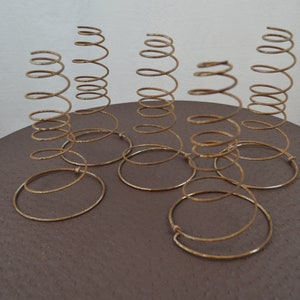 May include: Six gold metal springs, each with a circular base, arranged on a brown surface. The springs are stacked on top of each other, creating a decorative display.