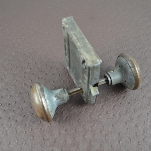 May include: An antique door knob set with a rectangular metal plate and two round knobs. The knobs and plate have a weathered, aged appearance with a mix of silver and bronze tones. The set is designed to be mounted on a door.