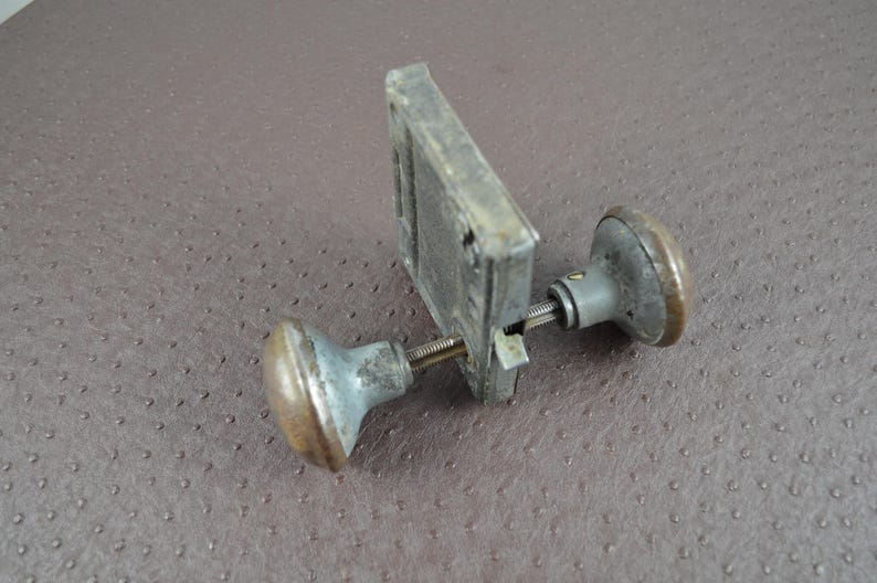 May include: An aged door knob set, featuring a rectangular metal plate and two circular knobs. The knobs and plate display a weathered, aged look with silver and bronze hues. The set is designed for door mounting.