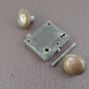 May include: An antique door lock set, comprising a rectangular lock body, two round knobs, and a threaded spindle. The metal components exhibit a weathered, aged appearance with a tarnished finish, indicative of vintage hardware.