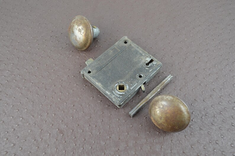 May include: An antique door lock set, comprising a rectangular lock body, two round knobs, and a threaded spindle. The metal components exhibit a weathered, aged appearance with a tarnished finish, indicative of vintage hardware.