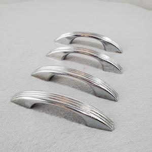 May include: A set of four silver-toned metal drawer pulls arranged in a diagonal line. Each handle features a curved, arched design with three distinct horizontal ridges running along the top surface and tapered ends for mounting purposes.