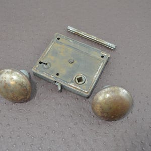 May include: A vintage door lock set, featuring two spherical knobs, a rectangular lock plate, and a threaded rod. The metal parts display a tarnished, aged look, indicative of antique hardware. This set is likely for a door or cabinet.