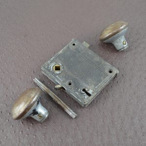 May include: An antique door lock set, comprising a rectangular metal plate, two oval doorknobs, and a connecting bar. The metal components have a weathered, aged appearance with silver and bronze tones, suggesting vintage hardware.
