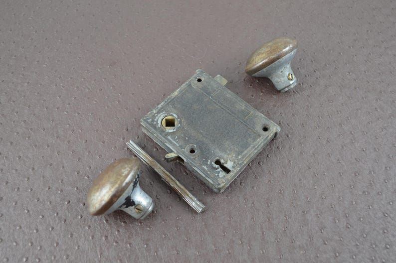 May include: An antique door lock set, comprising a rectangular metal plate, two oval doorknobs, and a connecting bar. The metal components have a weathered, aged appearance with silver and bronze tones, suggesting vintage hardware.