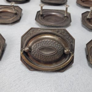 May include: Antique bronze drawer pulls with oval center designs and decorative floral patterns. Each pull has a handle and is attached with screws. The pulls are octagonal in shape and have a vintage aesthetic.