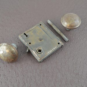 May include: An antique door lock set featuring two round knobs, a rectangular lock plate, and a connecting bar. The metal components have a tarnished, aged appearance, suggesting vintage hardware. The set is likely for a door or cabinet.