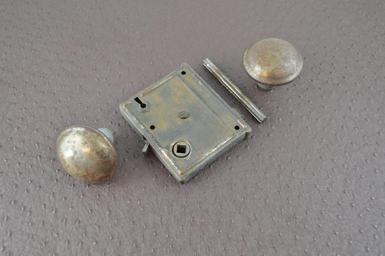 May include: An antique door lock set featuring two round knobs, a rectangular lock plate, and a connecting bar. The metal components have a tarnished, aged appearance, suggesting vintage hardware. The set is likely for a door or cabinet.